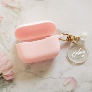 May include: A pink silicone case for wireless earbuds with a gold chain and a clear acrylic charm with the name "Amy" engraved on it.