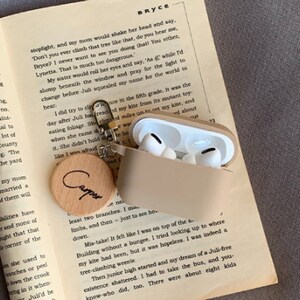 May include: A beige book with text on the pages. A brown wooden keychain with the word "Cooper" engraved on it is attached to a beige case for wireless earbuds. The case is open and the earbuds are visible.