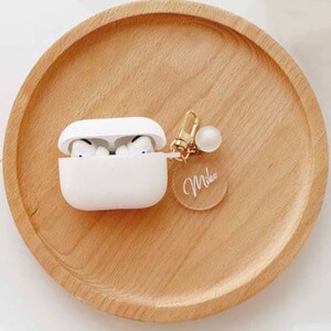 May include: White wireless headphone case with a gold chain and a clear charm with the name "Mika" written on it.