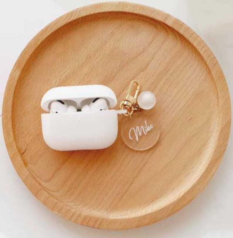 May include: White wireless headphone case with a gold chain and a clear charm with the name "Mika" written on it.