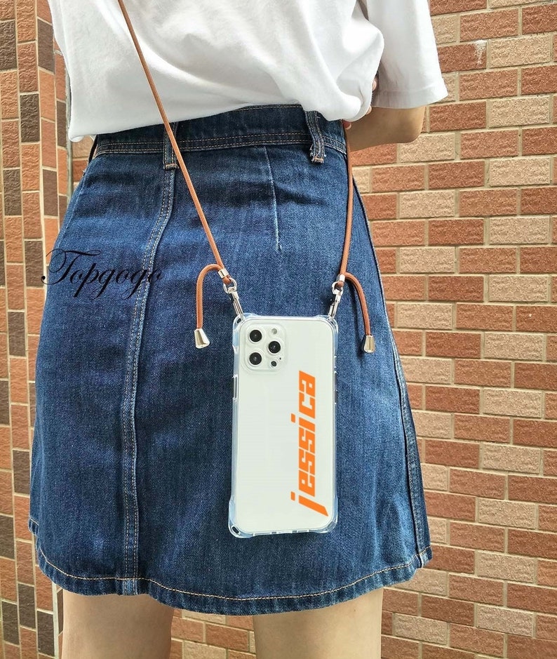 May include: A clear phone case with a brown leather phone strap. The case has a white background with the name "jessica" printed in orange letters.