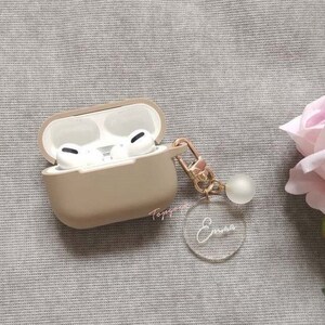 May include: A beige case for wireless earbuds with a clear acrylic keychain with the name "Emma" engraved on it. The keychain is attached to the case with a gold chain.
