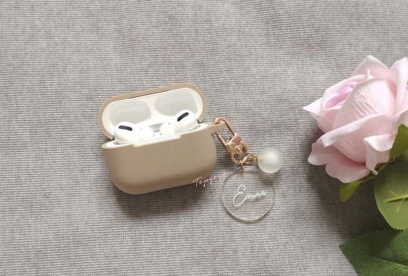 May include: A beige case for wireless earbuds with a clear acrylic keychain with the name "Emma" engraved on it. The keychain is attached to the case with a gold chain.