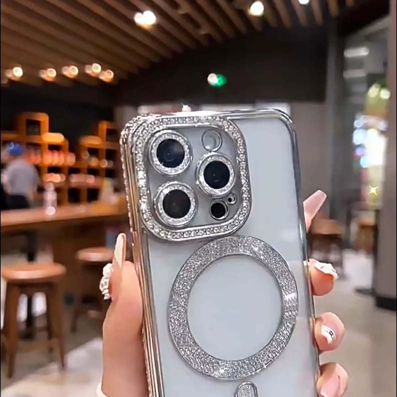 Op de afbeelding: Een transparante telefoonhoes met een zilveren rand en een grote ring van strass rond de camera lens. De hoes is versierd met strass.