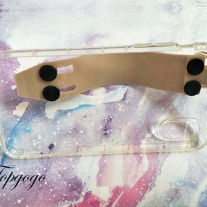 May include: A clear phone case with a beige silicone grip attached to the back. The grip has two black rubber pads on the bottom and one on the top.