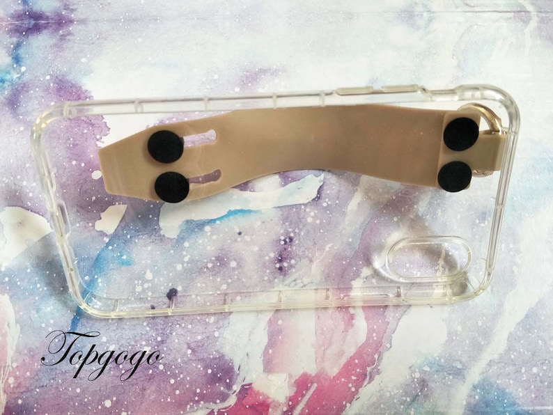 May include: A clear phone case with a beige silicone grip attached to the back. The grip has two black rubber pads on the bottom and one on the top.