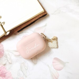 May include: A pink Airpods case with a white inscription that says "Sinda" and a pearl heart charm attached to a gold chain.