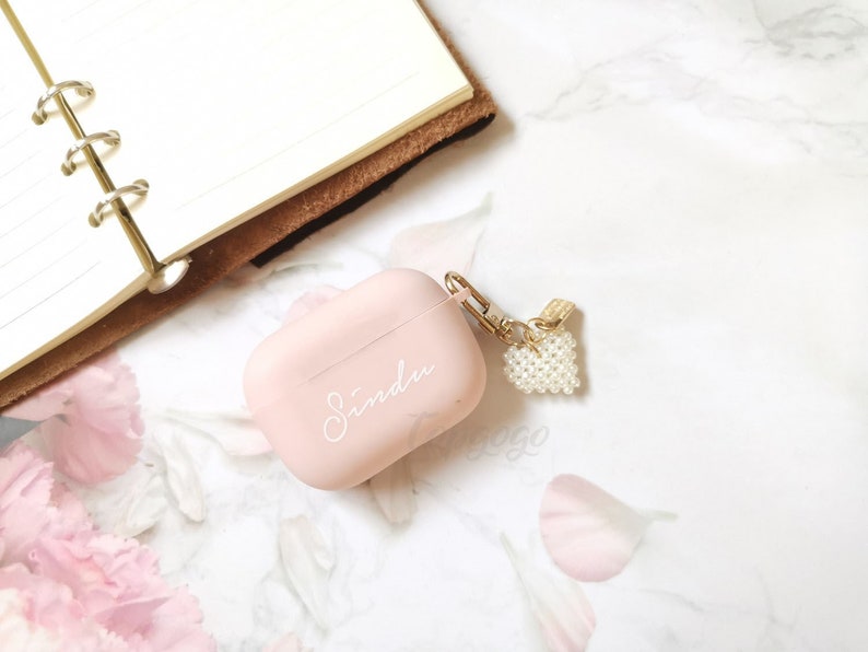 May include: A pink Airpods case with a white inscription that says "Sinda" and a pearl heart charm attached to a gold chain.