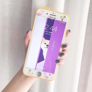 May include: A yellow phone case with a purple screen protector. The screen protector has a pink flamingo, palm trees, and a Shiba Inu with the text "なんだよ?" on it. The phone case also has a pineapple, a pink flamingo, and a heart sticker on it.