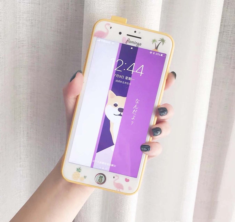 May include: A yellow phone case with a purple screen protector. The screen protector has a pink flamingo, palm trees, and a Shiba Inu with the text "なんだよ?" on it. The phone case also has a pineapple, a pink flamingo, and a heart sticker on it.
