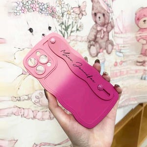 May include: A pink ombre phone case with a built-in phone grip and the name "Mini Jennifer" written on it.