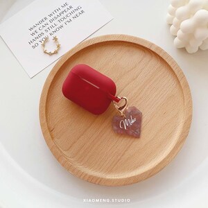 May include: Red silicone Airpods case with a gold chain and a pink heart charm with the word "Miko" engraved on it. The case is sitting on a wooden surface.