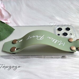 May include: A sage green phone grip with the name "Millie Perry" in white script. The grip is attached to a clear phone case with gold-colored rivets and a gold ring. The phone case is resting on a white, fluffy surface.