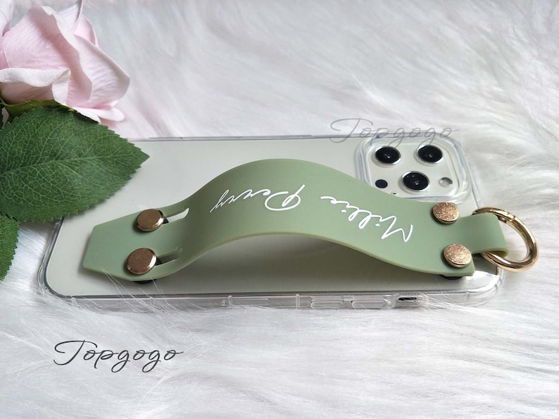 May include: A sage green phone grip with the name "Millie Perry" in white script. The grip is attached to a clear phone case with gold-colored rivets and a gold ring. The phone case is resting on a white, fluffy surface.
