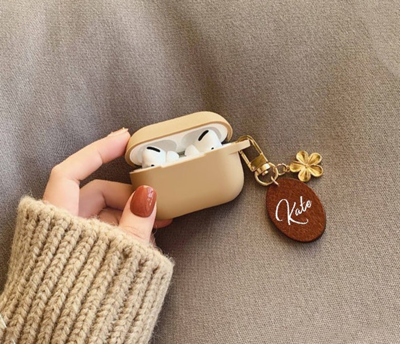 May include: A beige silicone case for AirPods with a gold chain and a brown leather keychain with the name "Kate" engraved on it.