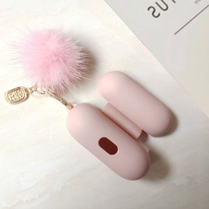 May include: A light pink AirPod case with a matching open lid and a fluffy pink pom-pom keychain. The keychain has a gold-colored clasp and a small rectangular charm. The case is designed to hold wireless earbuds.