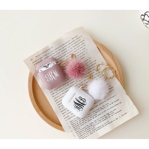 May include: Two personalised Airpod cases with fluffy keychains. One case is pink with the monogram "KM" and the other is white with the monogram "WB".