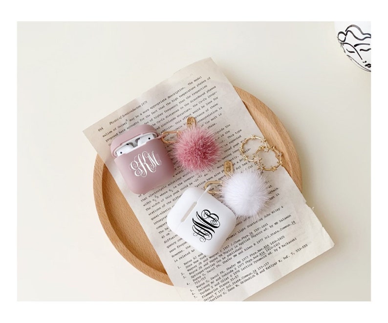 May include: Two personalised Airpod cases with fluffy keychains. One case is pink with the monogram "KM" and the other is white with the monogram "WB".