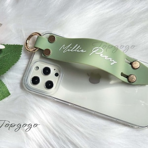 May include: Clear phone case with a light green strap featuring the name "Millie Perry" in white script. The strap has gold-colored metal accents and a gold ring. The phone case is on a white, fluffy surface.