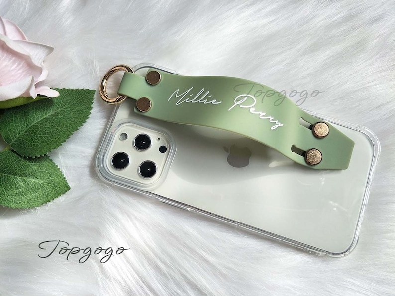 May include: Clear phone case with a light green strap featuring the name "Millie Perry" in white script. The strap has gold-colored metal accents and a gold ring. The phone case is on a white, fluffy surface.