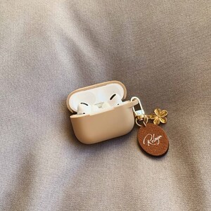 May include: A beige silicone case for wireless earbuds with a gold chain and a brown leather charm with the word "Robyn" written in white.