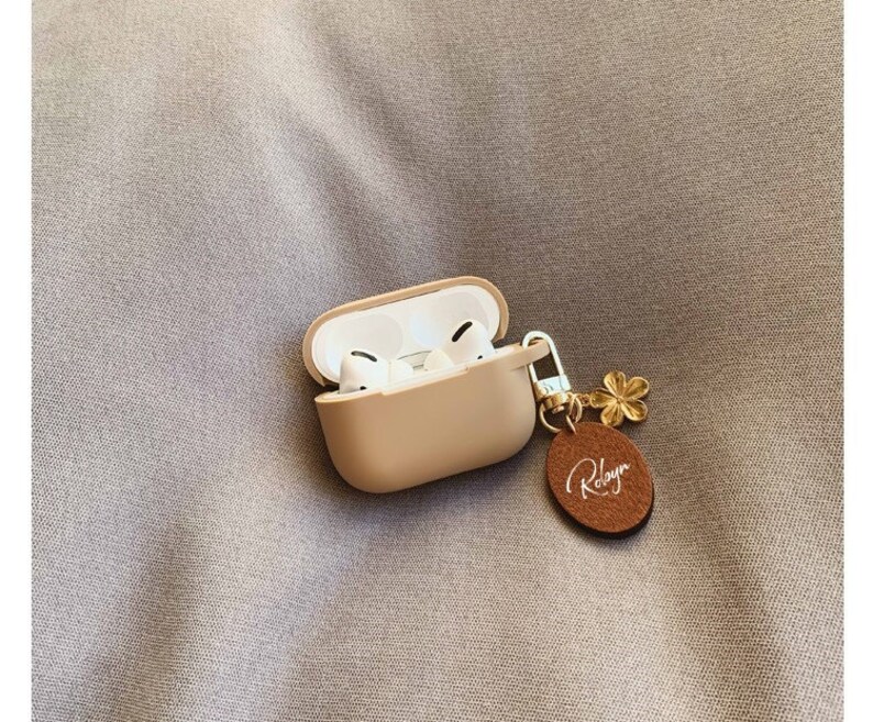 May include: A beige silicone case for wireless earbuds with a gold chain and a brown leather charm with the word "Robyn" written in white.