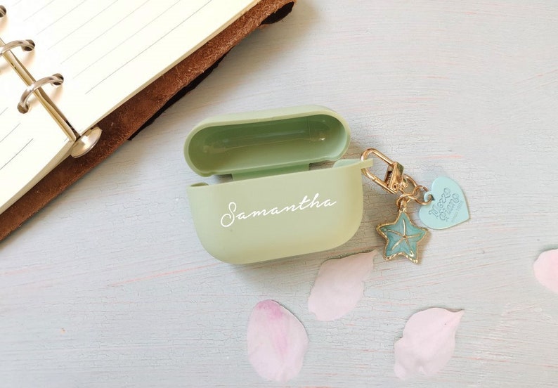 May include: A light green Airpods case with the name Samantha written in white on the front. The case has a gold charm attached to it with a blue starfish and a heart that says "More Than Words".