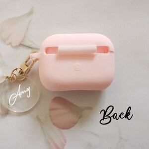 May include: A pink silicone case for AirPods with a gold chain and a clear charm with the name "Amy" engraved on it. The case has a small opening for the charging port. The back of the case is shown in the image.