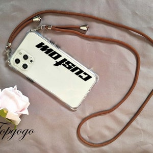 May include: A clear phone case with a brown phone strap. The case has the word "Custom" printed on it in black.