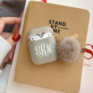May include: A grey Airpods case with a white monogram "KM" and a fluffy grey pom-pom keychain. The case is sitting on a brown notebook with the text "Stand by Me The Journal" printed on the cover.