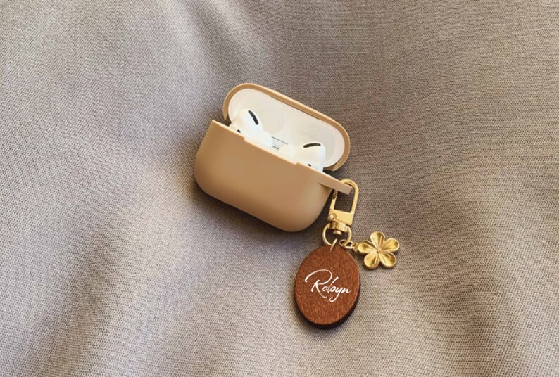 May include: A beige wireless earbud case with a gold keychain featuring a small flower charm and a brown oval tag with the name "Robyn" written in white.