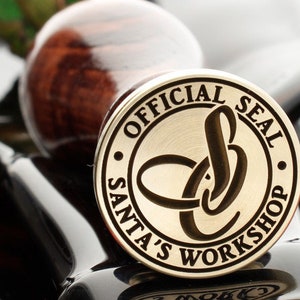 Könnte beinhalten: Ein goldfarbenes Metallstempel mit dem Text "Official Seal Santa's Workshop" am Rand. Der Stempel hat ein stilisiertes Design in der Mitte.