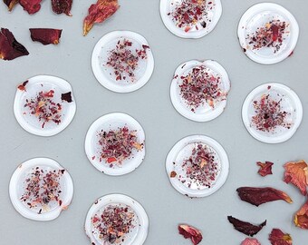 Peut inclure: Douze contenants ronds blancs avec un dessus transparent, chacun rempli de pétales de rose séchées. Les contenants sont disposés en grille sur une surface gris clair.