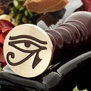 May include: A brass wax seal stamp featuring a detailed design of the Eye of Horus, an ancient Egyptian symbol of protection and power. The stamp is mounted on a dark red wooden handle.