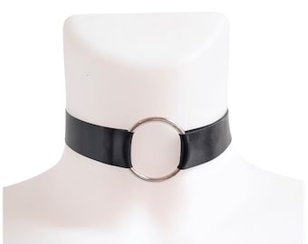 May include: A black choker necklace with a silver-colored metal ring in the center. The choker is displayed on a white mannequin head. The necklace is a simple, yet bold accessory.