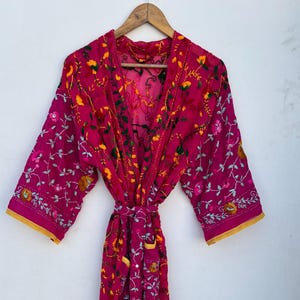 May include: A magenta-colored robe with floral embroidery in yellow, green, and white. The robe has a tie closure and yellow trim on the sleeves and edges. It is hanging on a wooden hanger.