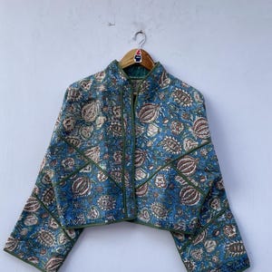 以下が含まれることがあります： 青い花柄のジャケット。ハイカラーとジッパー開閉。茶色と緑のアクセントが施されたパッチワークデザイン。袖は七分丈。木製のハンガーにかかっています。