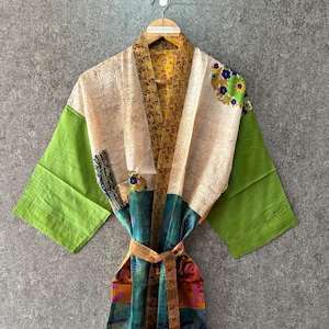 May include: A colorful kimono-style robe with a patterned design. The robe features a mix of colors, including green sleeves, cream-colored lapels, and a patterned interior. A brown belt cinches the waist. The robe is hanging on a wooden hanger.