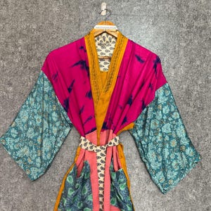May include: A colorful kimono-style robe with a mix of patterns and colors. The robe features a vibrant pink body, teal sleeves with a floral design, and a mustard yellow trim. It includes a matching belt and pockets.