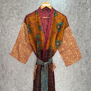 May include: A colorful, patterned robe with a belt. The robe features a mix of floral and geometric designs in shades of gold, red, and blue. The robe is hanging on a wooden hanger against a gray background.