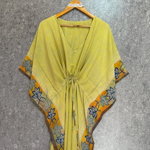 Puede incluir: Vestido caftán amarillo con cuello en V y cintura con cordón. El vestido presenta un borde floral en azul, naranja y negro. El caftán está colgado de una percha de madera.