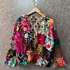 Puede incluir: Una chaqueta de manga larga con un diseño de patchwork hecho de varias telas bordadas coloridas. El exterior muestra diversos patrones florales y geométricos en tonos rojo, rosa, amarillo y azul. El forro interior tiene un patrón de rayas marrones.