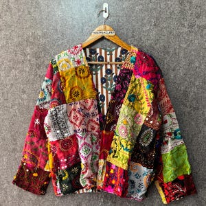 Könnte beinhalten: Eine langärmelige Jacke mit einem Patchwork-Design aus verschiedenen bunten Stoffen mit aufwendigen Stickereien und kleinen Spiegelakzenten. Das Innenfutter zeigt ein braun-weißes vertikales Streifenmuster mit blauen Blumenmotiven.