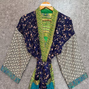 Op de afbeelding: Een marineblauwe kimono-jurk met bloemenprint en groene en turquoise accenten. De jurk heeft een striksluiting, mouwen met patronen en zakken. De jurk hangt aan een houten hanger.