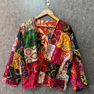 Könnte beinhalten: Eine langärmelige Jacke mit einem lebendigen Patchwork-Design und verschiedenen gestickten kreisförmigen Mustern in Rot, Gelb, Grün und Weiß. Das Kleidungsstück hat einen V-Ausschnitt und hängt an einem Holzkleiderbügel mit dem Etikett HASTHSHILPAM.