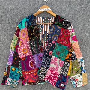Könnte beinhalten: Eine langärmelige Jacke mit einem lebendigen Patchwork-Design aus verschiedenen bestickten Stoffen in Farben wie Rot, Pink, Lila, Türkis und Schwarz. Das Innenfutter zeigt ein braun-weißes vertikales Streifenmuster mit floralen Motiven.