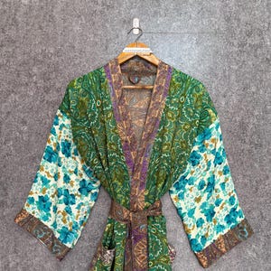 Puede incluir: Bata kimono verde con estampado de cachemira y mangas florales turquesas y blancas. La bata tiene un cinturón a juego y se exhibe en una percha de madera. El ribete tiene un patrón marrón y morado en contraste.