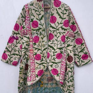 May include: Cream-colored embroidered jacket with a floral pattern in pink and green. The jacket features a long, open design with a contrasting patterned lining. The embroidery includes stylized flowers and vines, creating a bohemian aesthetic. The jacket is a unique piece of apparel.