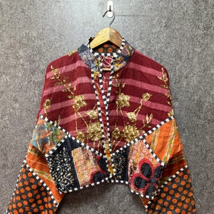 Op de afbeelding: Een kort jasje met een patchworkontwerp, een bordeauxrood gestreept bovendeel, gouden bloemenborduursels en onderste panelen met oranje, zwarte en witte patronen. Het kledingstuk heeft wijde mouwen en een zwart-wit geblokte bies.