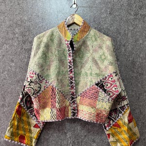 May include: A cropped jacket featuring a patchwork design of various fabrics in shades of green, pink, yellow, and black. The garment has a mandarin collar, long sleeves, and a front opening with a dark patterned trim and visible running stitch details.